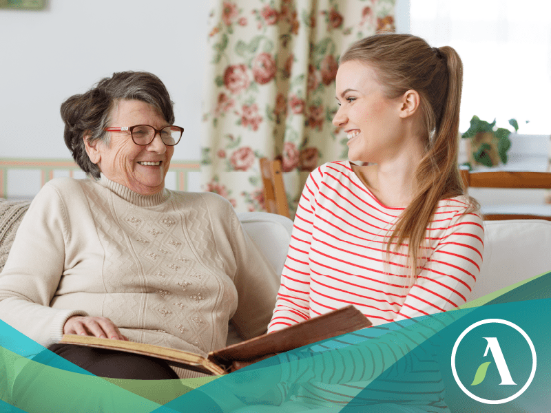 Grandma and teenage granddaughter sit on a couch in a sunny living room, smiling and looking at a photo album.