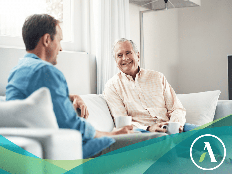 Two white men, one aged 40, one aged 70, sit on couch with coffee and chat.