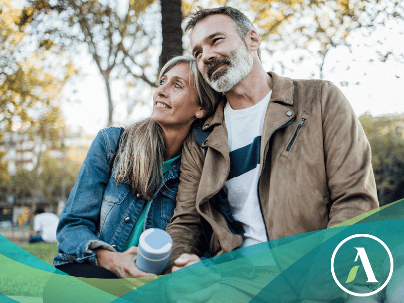 Caucasian couple hold hands on a bench, smiling and looking off into the distance on a fall or spring day.